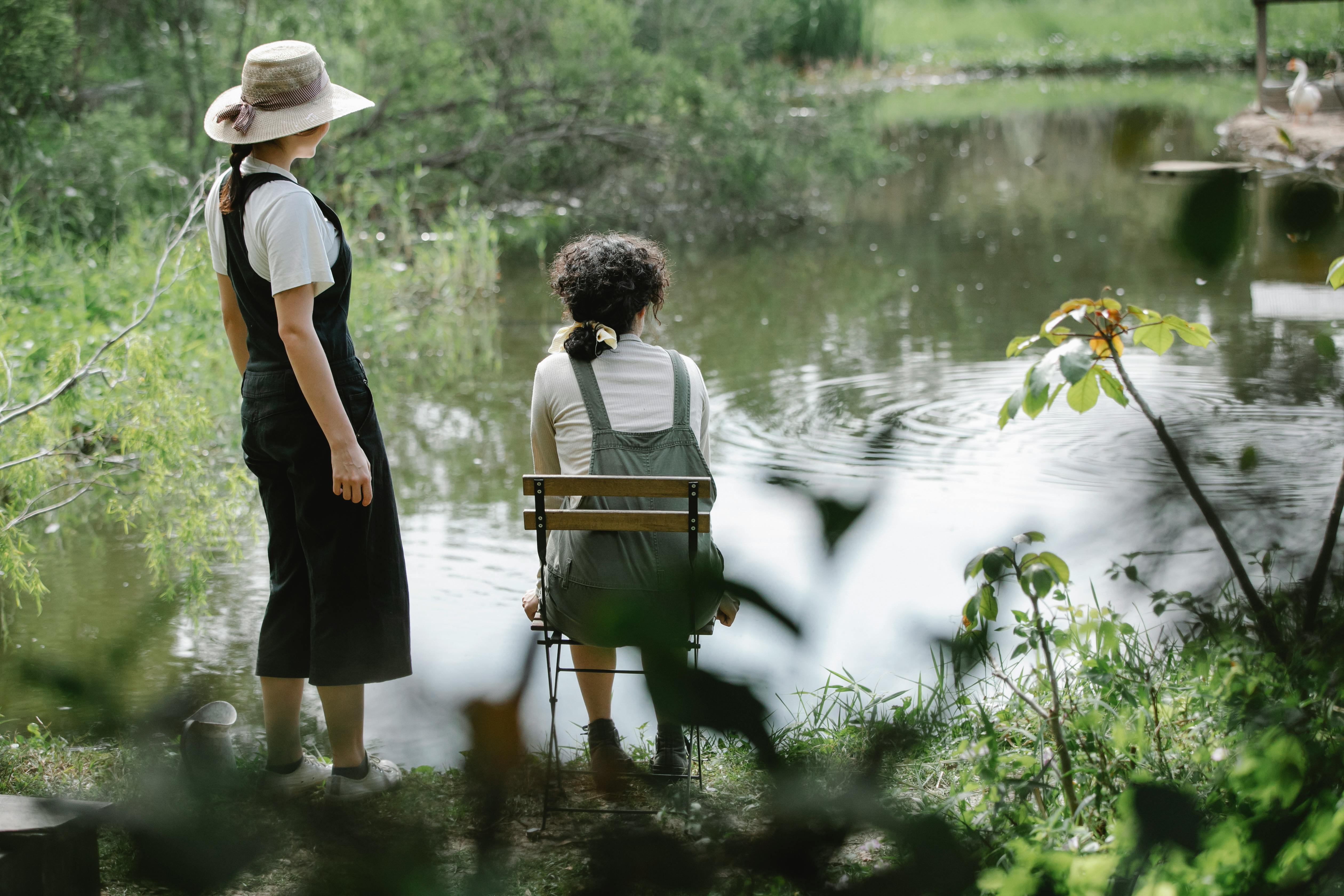 Back view full body of unrecognizable females in overalls resting on  coast of pond in village