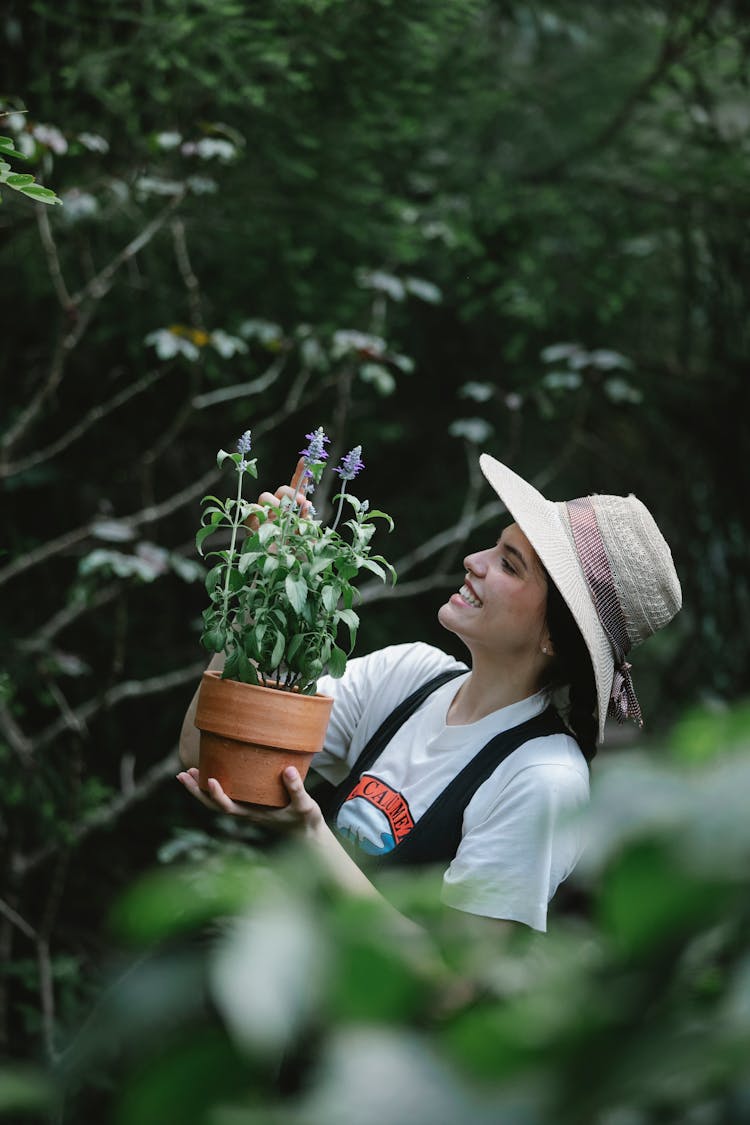 Positive Female Gardener With Potted Salvia In Garden