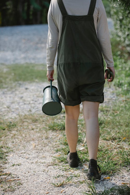 Back view of anonymous female gardener walking on pathway with watering can in farm