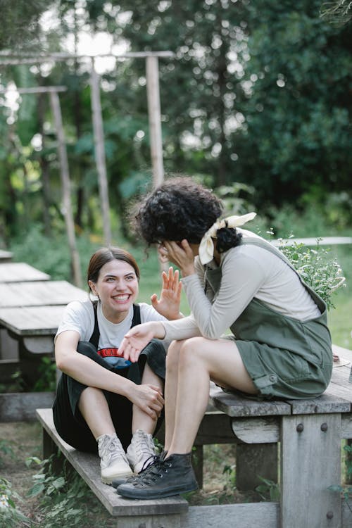 Full length of smiling young women in overalls having conversation while sitting on wooden bench in camp near green trees and grass with plants and looking at each other