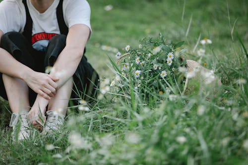 Crop faceless female in overall and t shirt sitting on green field with grass and white flowers near plants in summer day in nature
