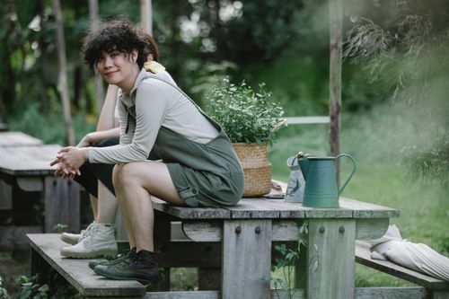 Free Full length side view of multiethnic young ladies in overalls sitting on wooden bench near basket with wildflowers and pots in camp near green grass and forest in summer day Stock Photo