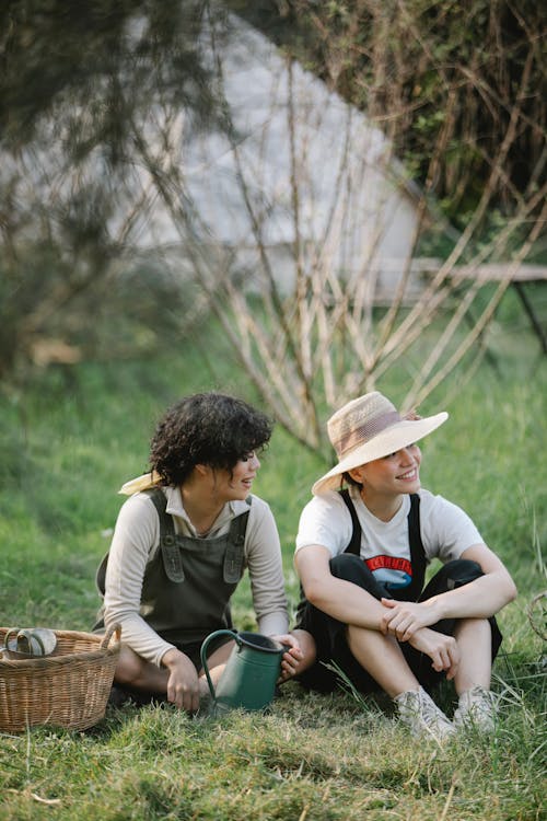 Full body of smiling young female gardeners in overalls and straw hats sitting on green meadow in nature near trees and basket with pot in summer day and looking away