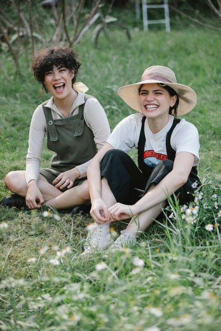 Full length of smiling multiracial young gardeners in overalls and straw hats sitting on grassy field in nature near flowers and plants with trees in summer while looking at camera