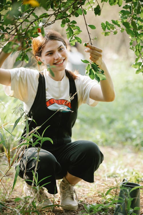 Full length of smiling young woman in overall touching tree branch with green leaves near pot on grassy meadow in sunny summer day in nature