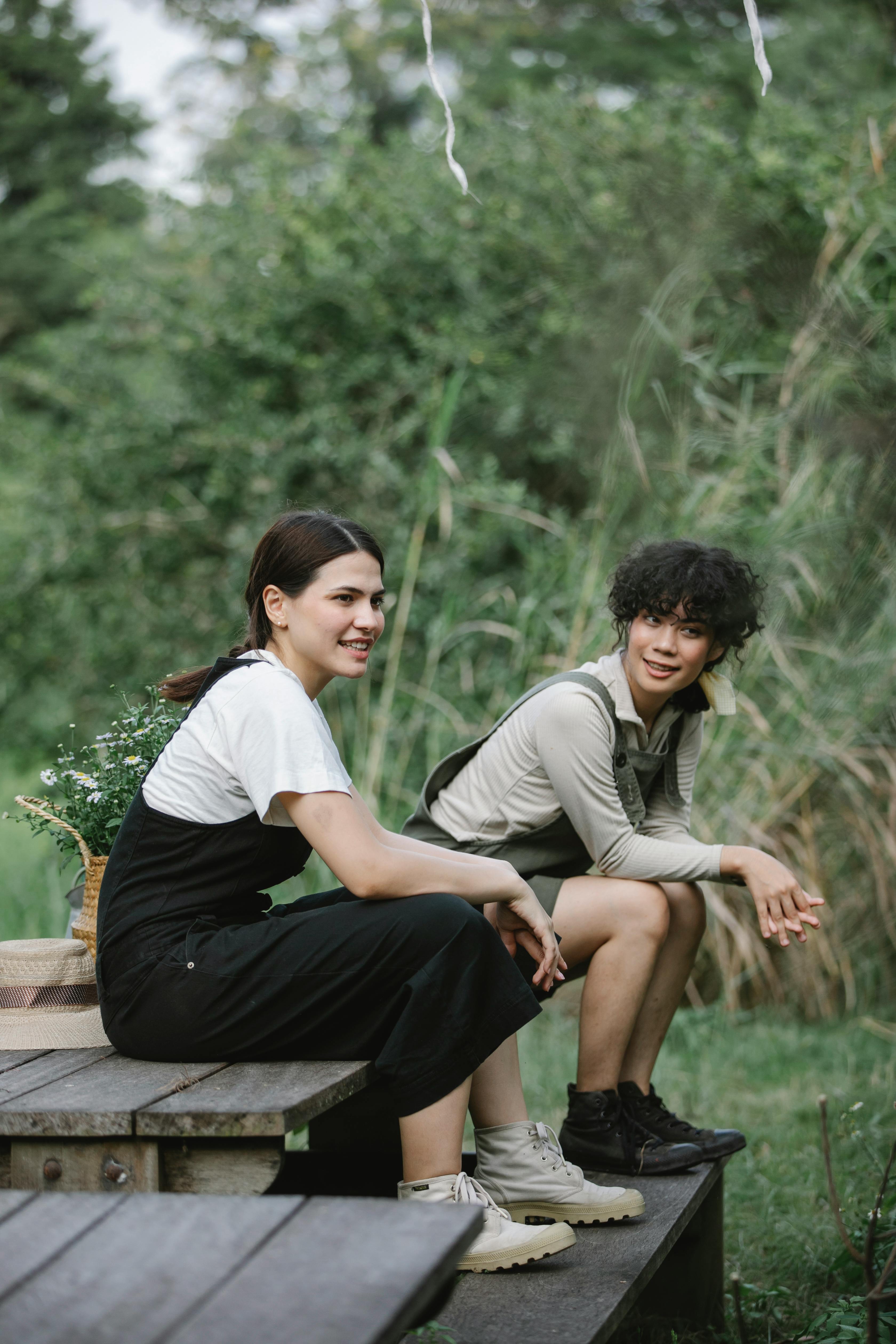 Full body side view of multiracial young women in overalls sitting on wooden bench near basket with wildflowers in park near green grass and bushes with forest in summer day