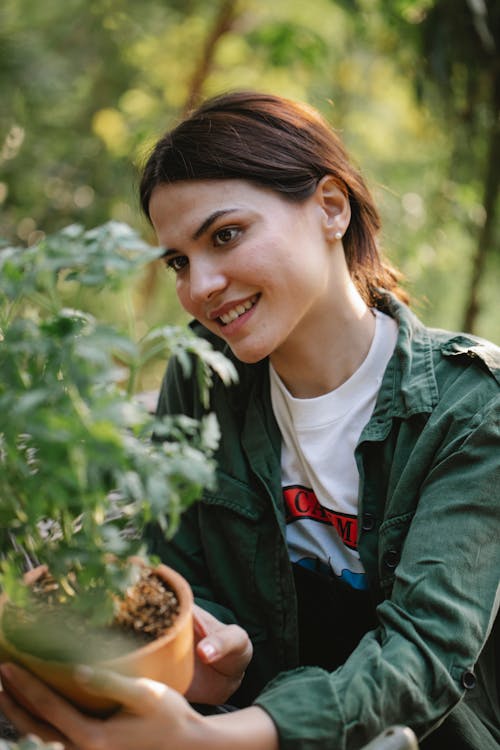 Free Cheerful sincere young female grower looking at green plant in pot on sunny day Stock Photo
