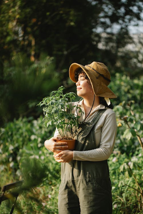 Content ethnic female grower in panama hat with potted plant and closed eyes in summer garden