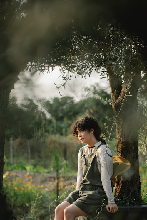 Dreamy ethnic female horticulturist sitting on pot with olive tree while looking away on farmland