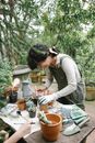 Side view of concentrated ethnic female gardener standing at wooden table with gardening tools while planting flower in pots during work in orangery