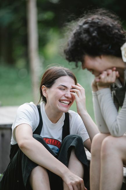 Positive ethnic female coworkers in uniform laughing while sitting on wooden bench during work break in garden on blurred background