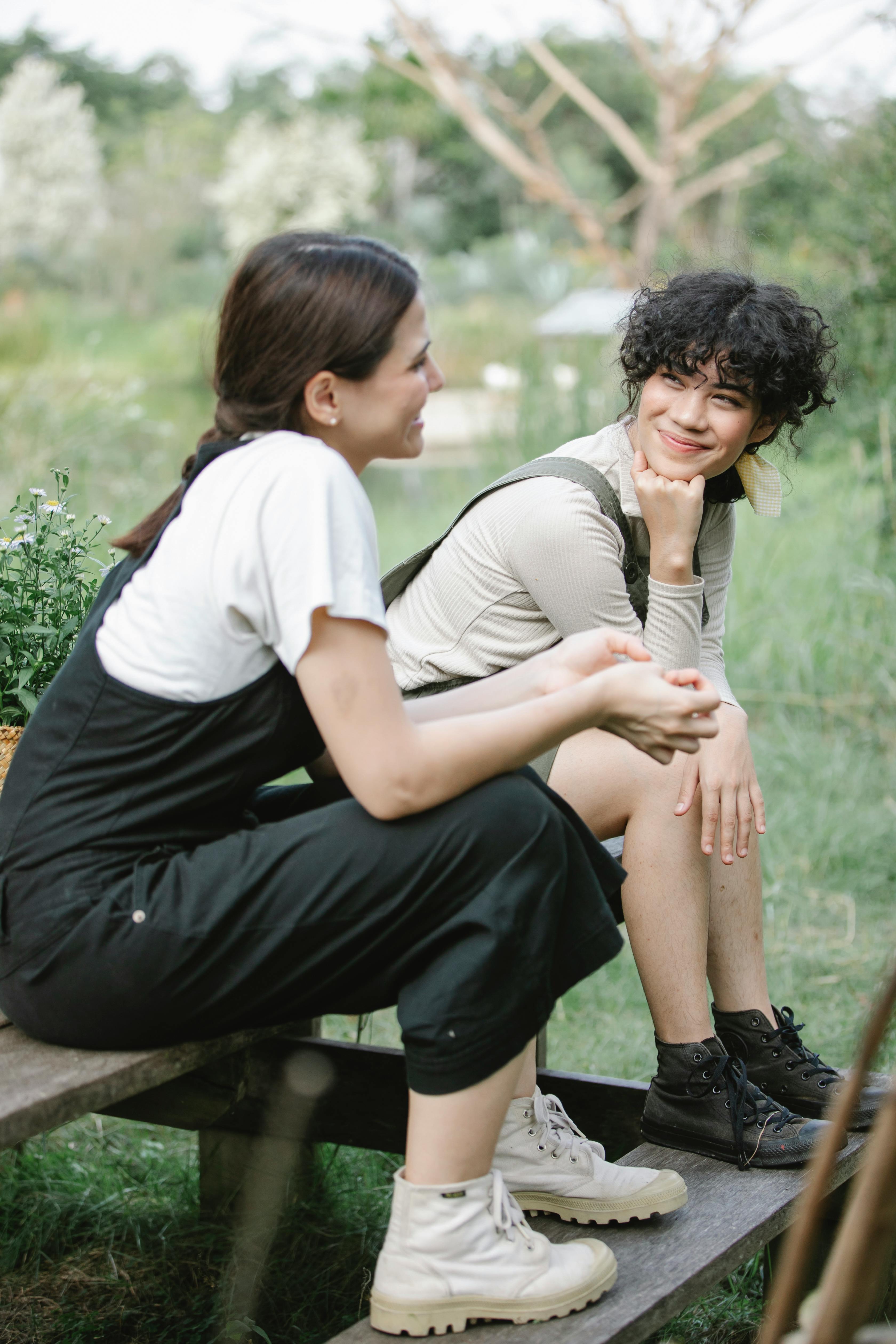 Two women in casual attire converse on a wooden bench in a lush garden setting.