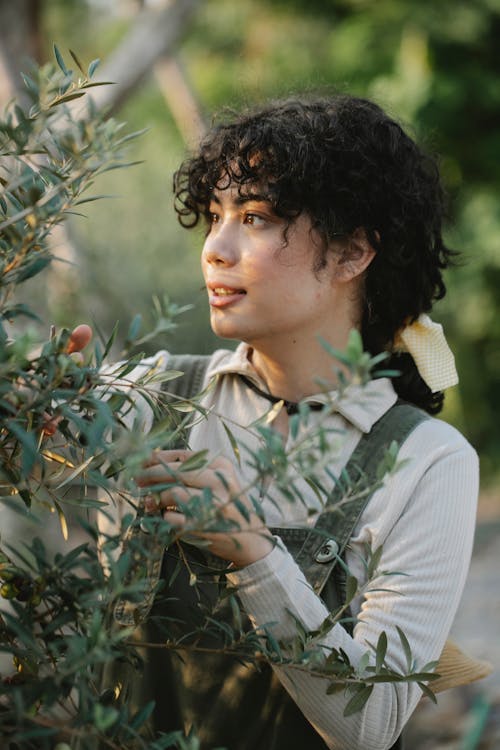 Free Side view of ethnic female gardener in uniform standing near sprigs of blooming tree while working in garden on blurred background Stock Photo