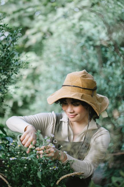 Positive ethnic female gardener in straw hat with scissors cutting stems of small flowers in garden with green plants on blurred background