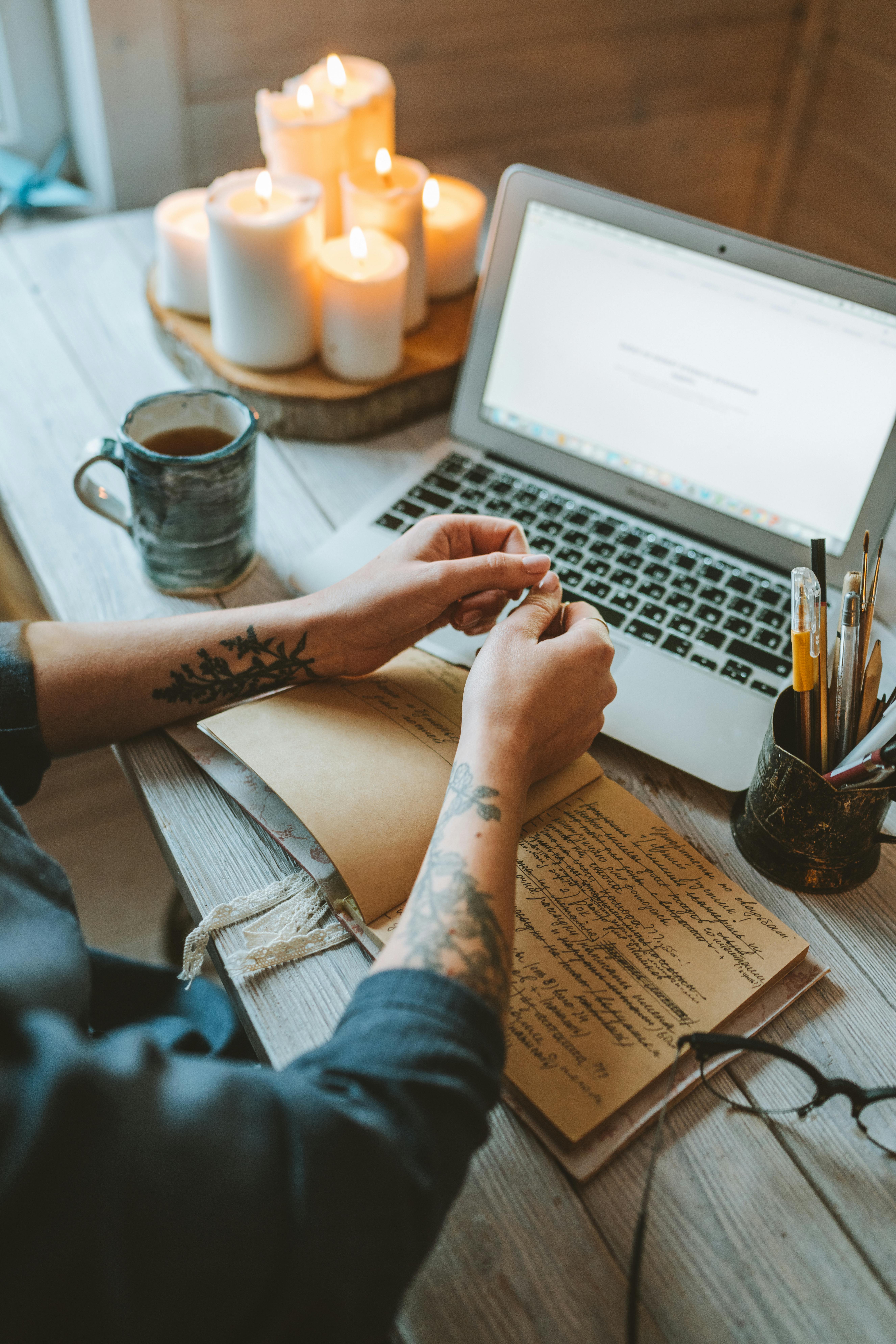 A warm, inviting workspace featuring a laptop, candles, and a person writing notes.