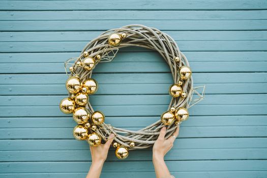 Hand holding an elegant Christmas wreath decorated with golden ornaments against a blue wooden background.