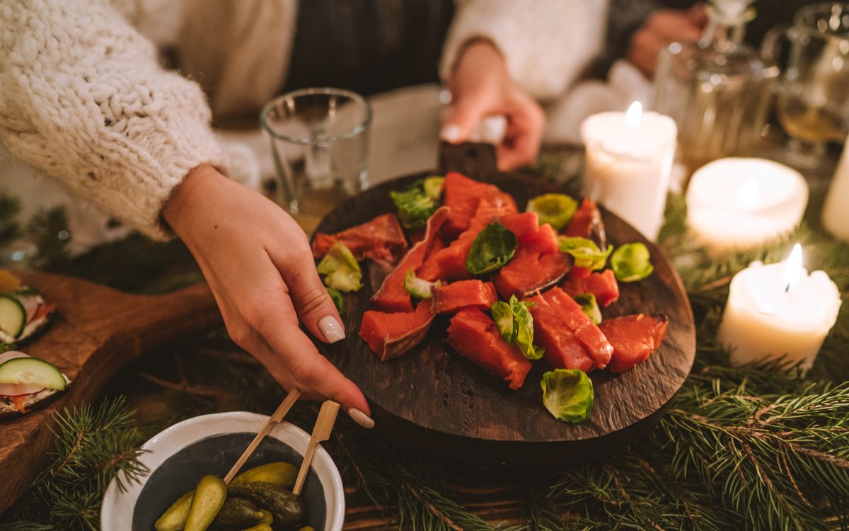 A cozy holiday table setting featuring smoked salmon, candles, pickles, and greenery.