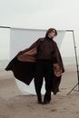 Melancholy man model in trendy clothes standing in raised hand in pocket on sandy  beach near ocean against white fabric background in cloudy day