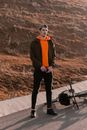 Man in Brown Jacket and Orange Backpack Standing on Road