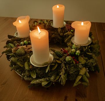 From above white thick candlesticks burning on creative wreath holder made of artificial plant leaves and placed on wooden table in dark room