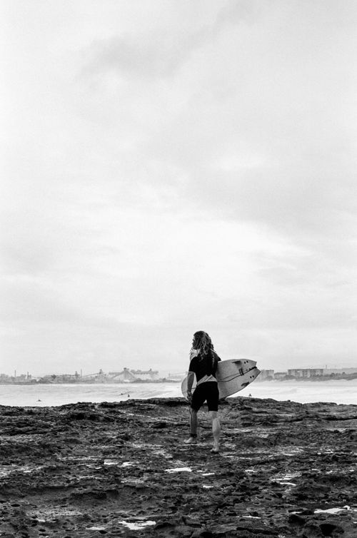 Free A surfer holding a surfboard walks on a rocky shore on a cloudy day. Stock Photo