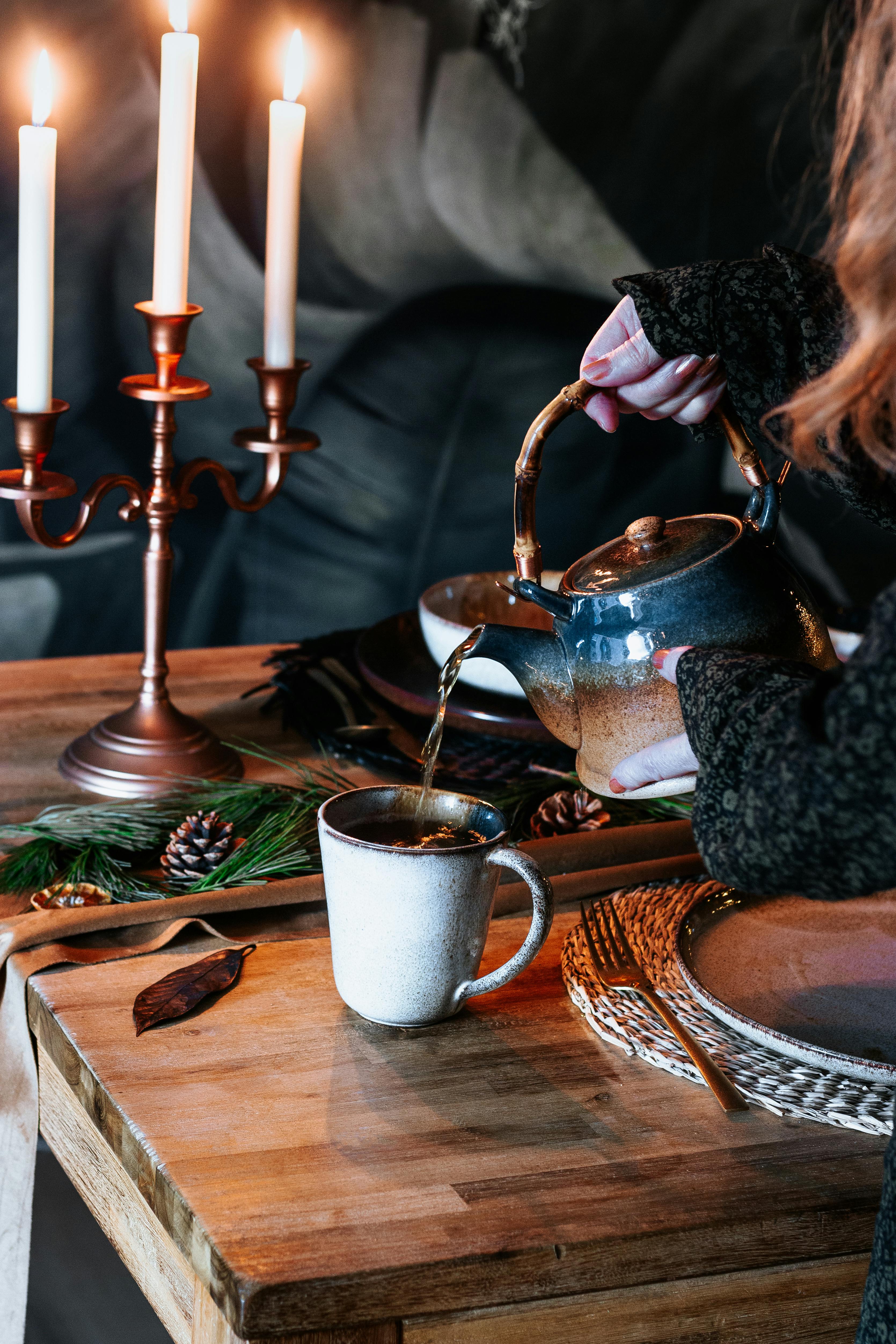 A warm and inviting image of tea being poured from a teapot into a cup surrounded by burning candles on a wooden table.