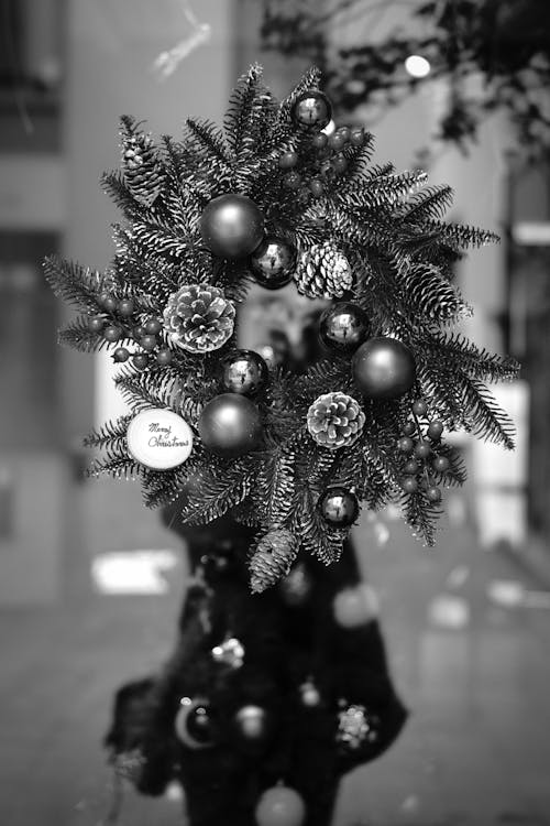 Black and white of Christmas wreath decorated with small shiny baubles and pine cones hanging on door of modern building during festive event