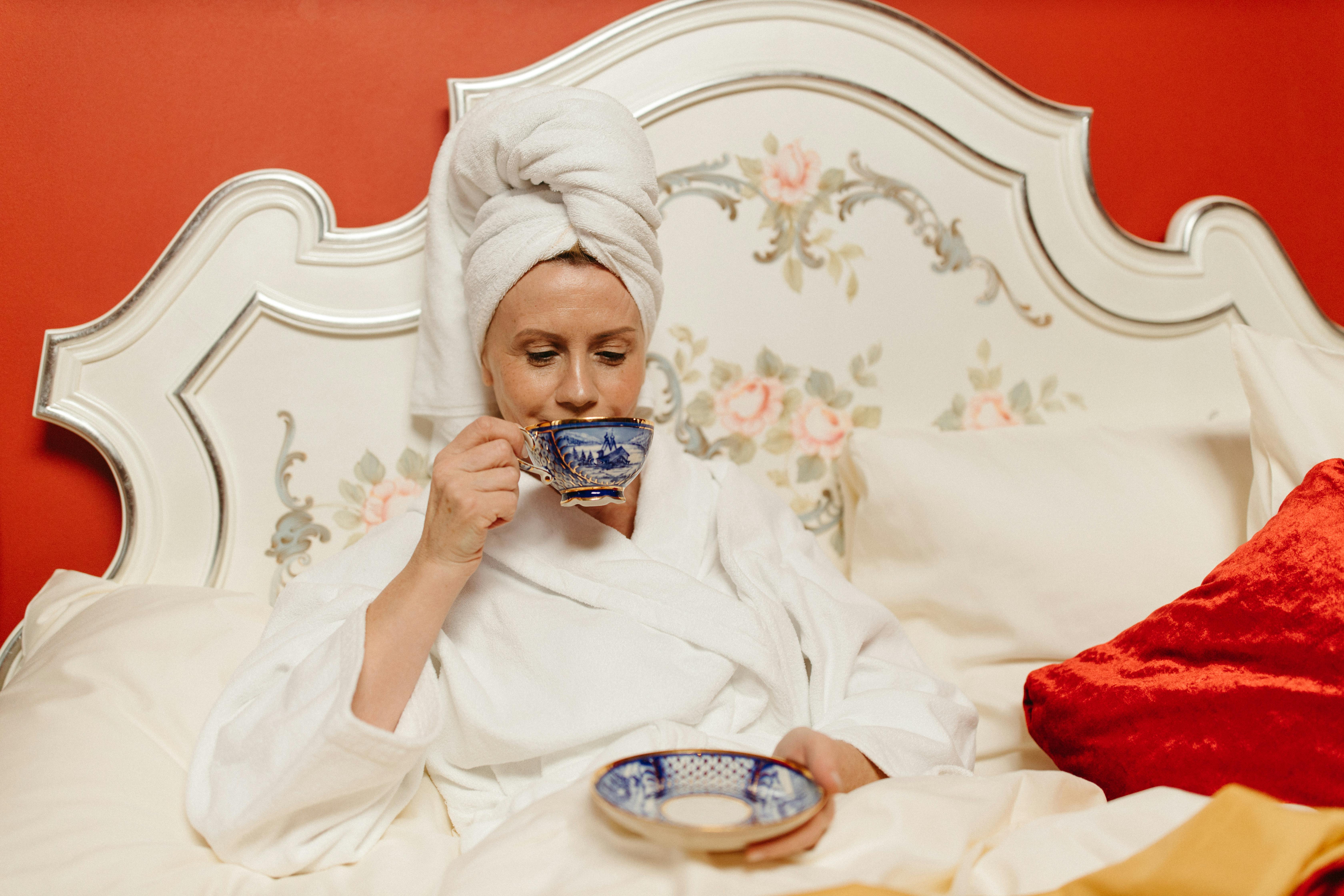 A woman in a white robe and towel wrap enjoys tea in a cozy bedroom setting.