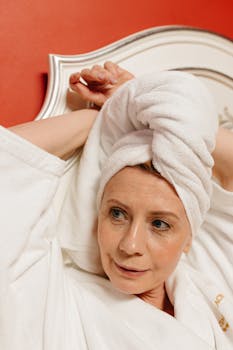 Middle-aged woman in bathrobe and head towel, relaxing indoors against a red background.