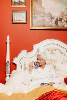 Woman in bathrobe with head towel sipping tea in an elegant bedroom.