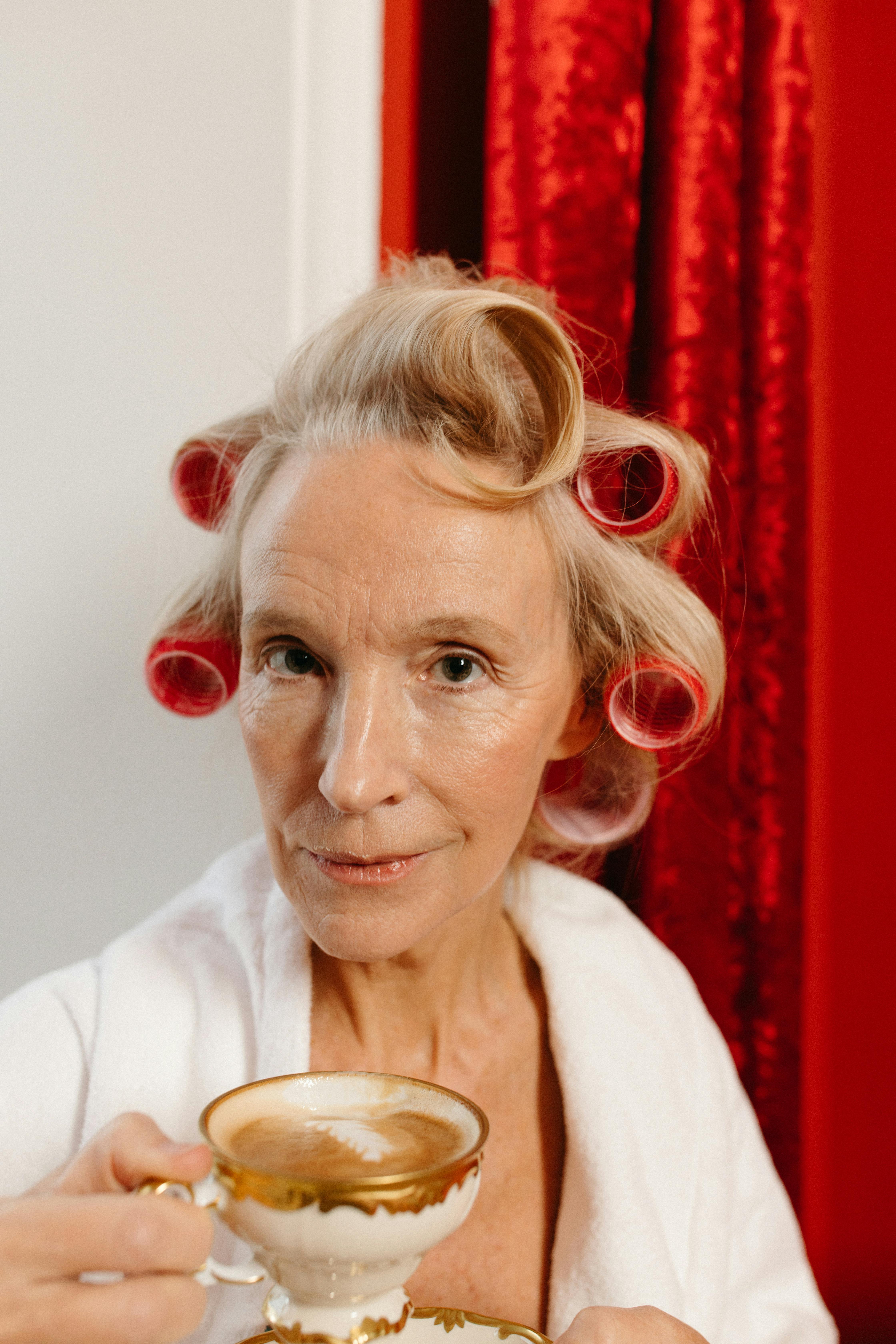 A sophisticated elderly woman with hair rollers enjoying a cup of coffee indoors.