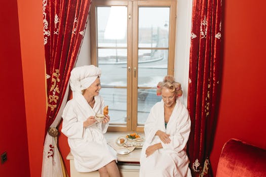 Two senior women in bathrobes enjoy breakfast by a window in a cozy hotel room.