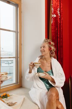 Woman in bathrobe enjoying coffee by the window with hair rollers in. Relaxing morning vibes.