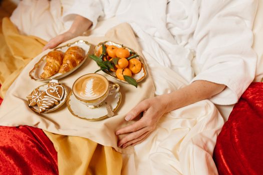A comforting breakfast setup with latte, croissants, gingerbread, and mandarins on a bed tray.