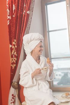 Woman enjoying a coffee and croissant by the window in a bathrobe at home.
