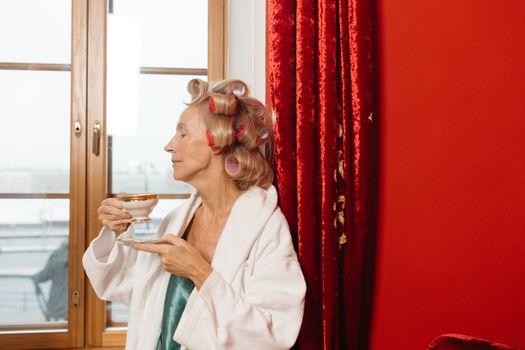Relaxed senior woman in bathrobe with curlers, savoring tea by the window.