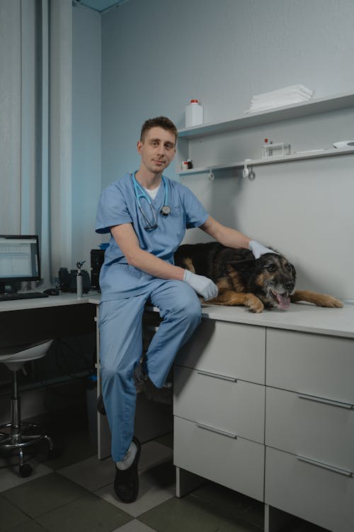 Man in Blue Scrub Suit Sitting on Table While Holding a Dog