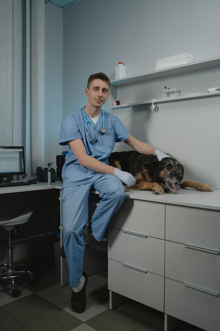 Man In Blue Scrub Suit Sitting On Table While Holding A Dog