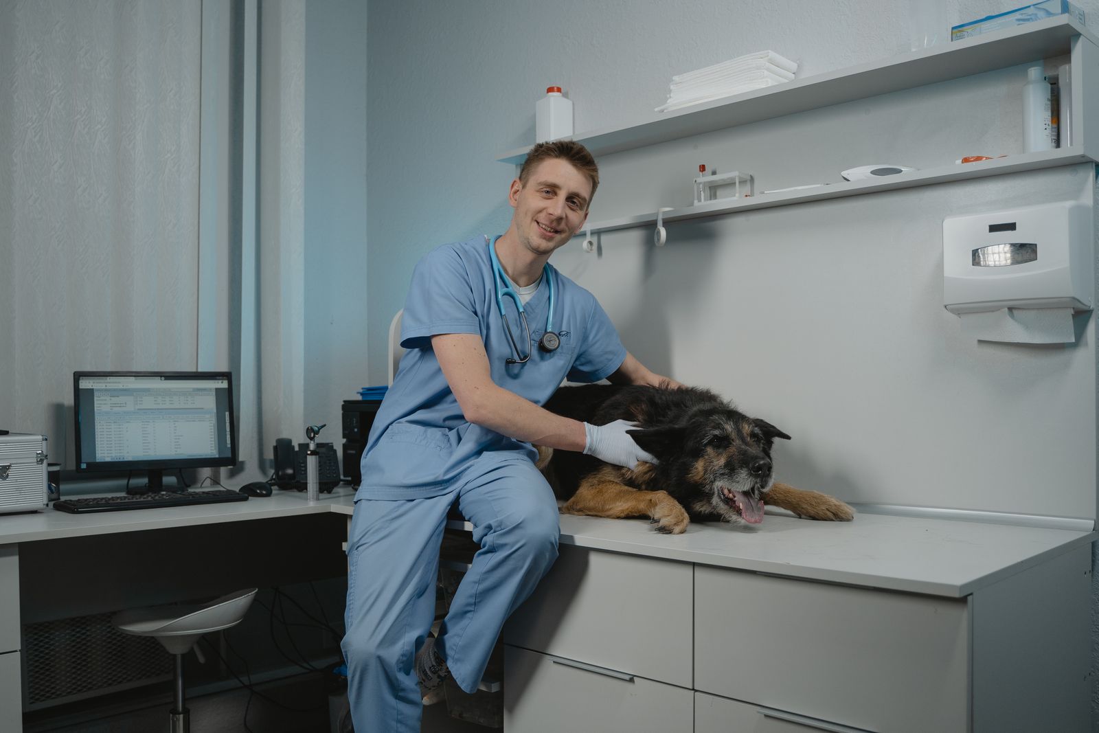 A smiling vet in blue scrubs sitting beside an older German Shepherd on the examination table