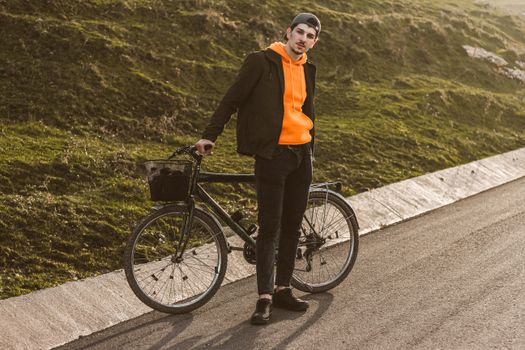 A stylish man leans on his bicycle on a sunlit road, embracing leisure and exploration.