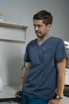 Tattooed male healthcare worker in scrubs with stethoscope, standing in a clinic.