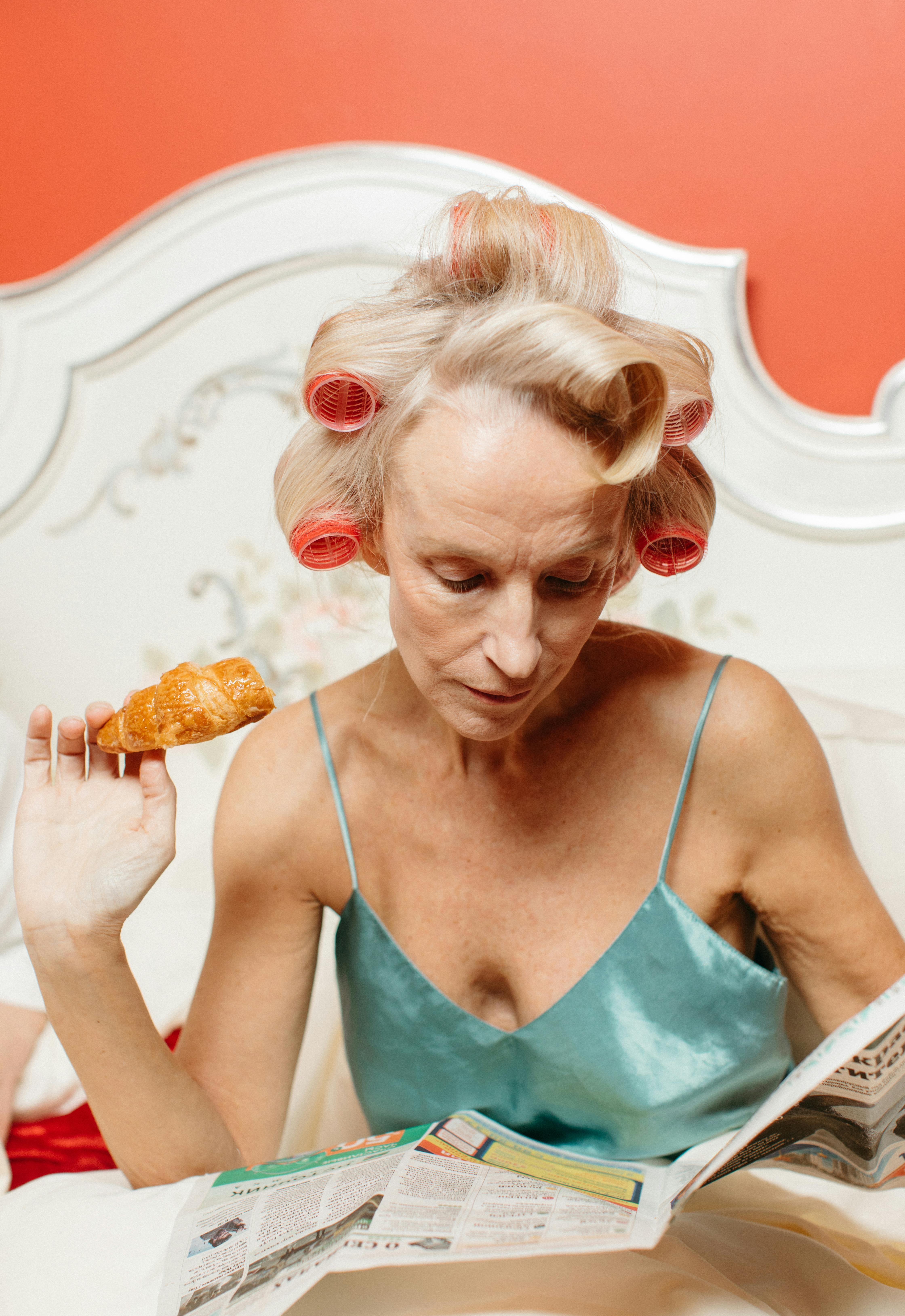 Woman in rollers enjoys a croissant while reading a newspaper in bed, wearing a nightgown.