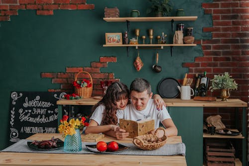 Free Positive Asian couple embracing while reading recipe book together sitting at table in kitchen Stock Photo