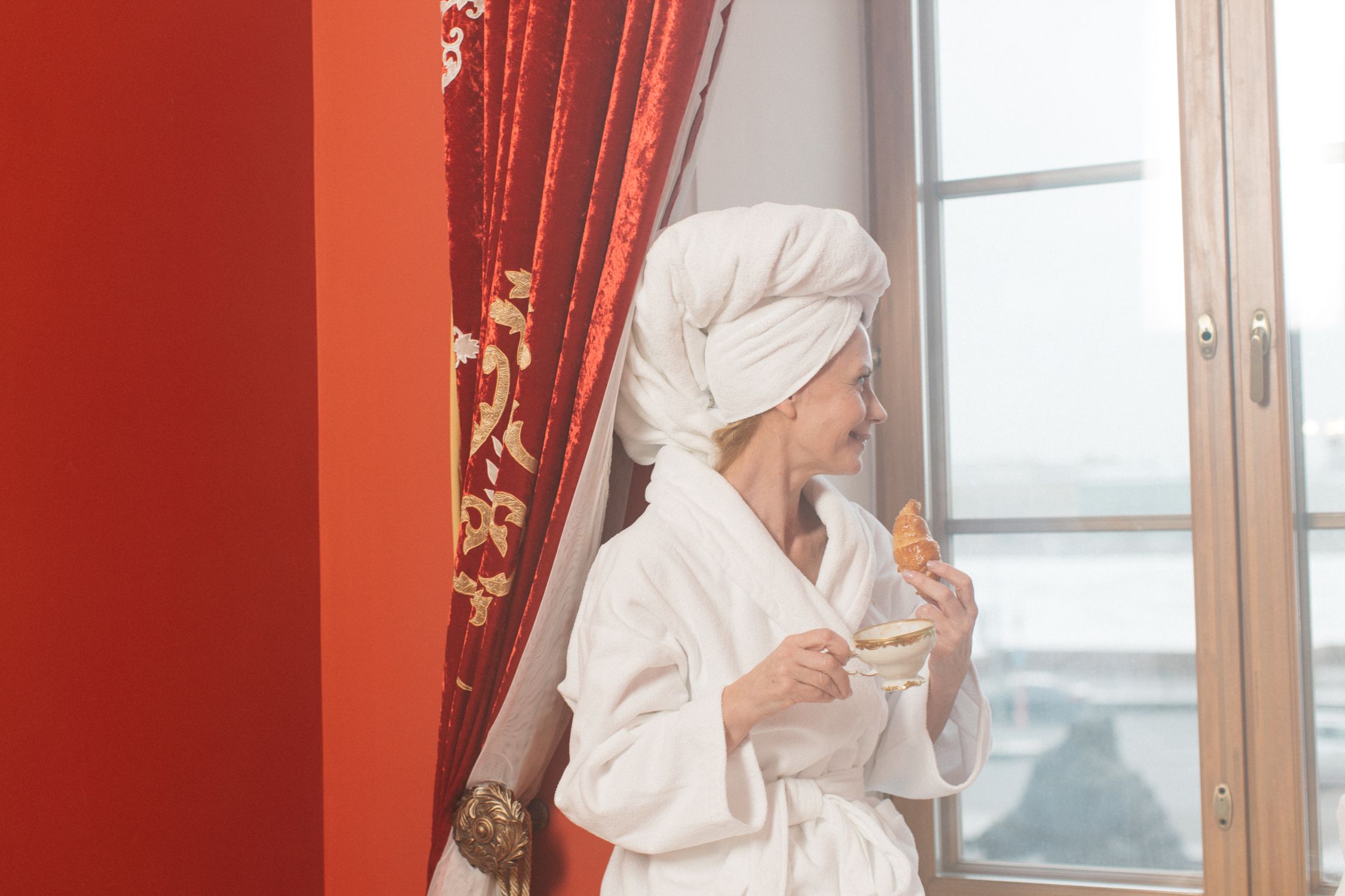 Woman in bathrobe with coffee and croissant looks out window, enjoying a leisurely morning.