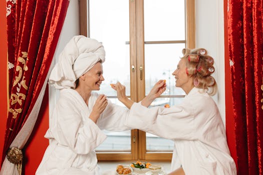 Two women in bathrobes enjoying a relaxing spa day indoors, sharing food and laughter by the window.