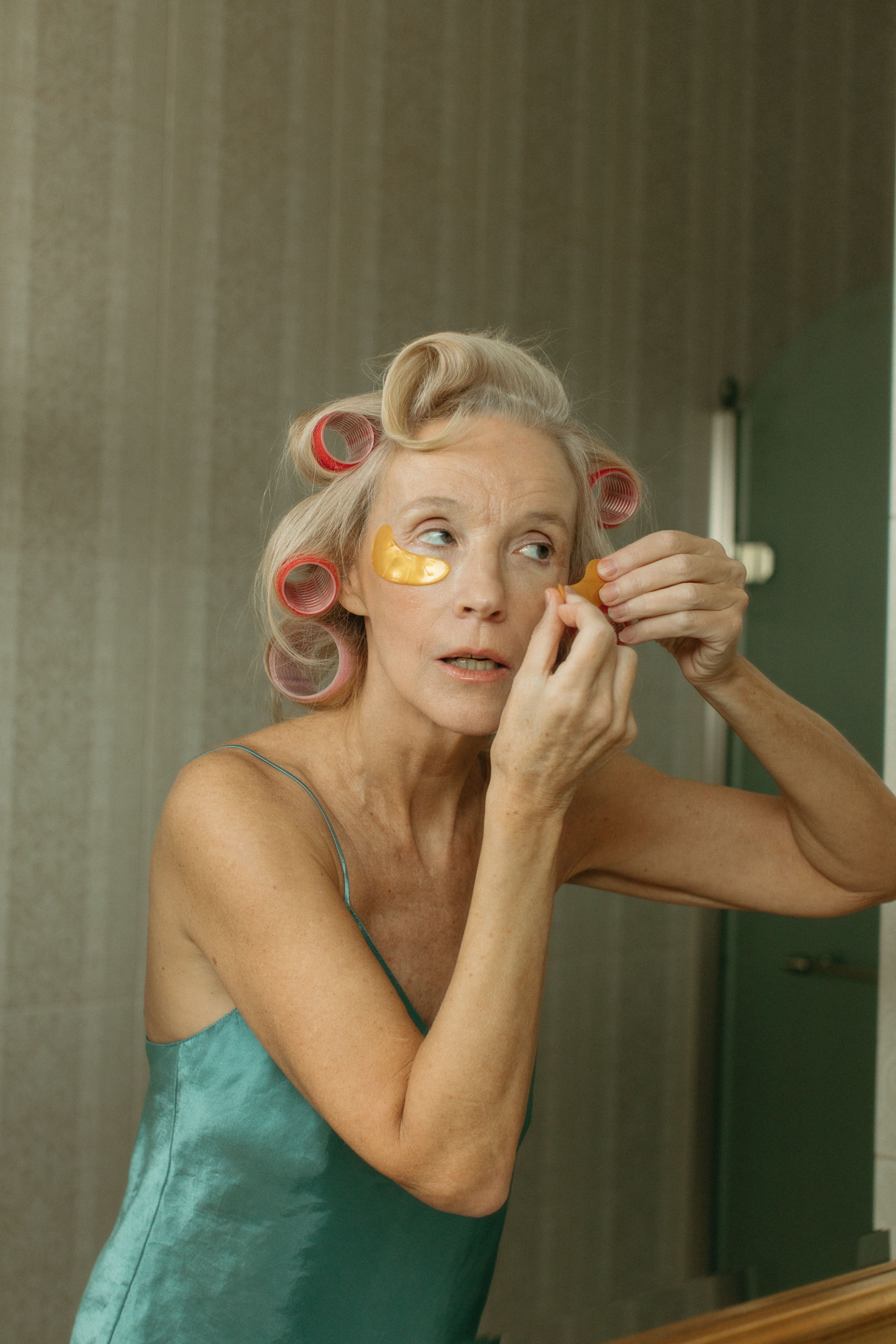 Elderly woman applies skincare patches with hair in curlers indoors.