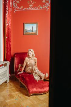 A woman in an elegant dress sits gracefully on a red chaise lounge in a beautifully decorated room.