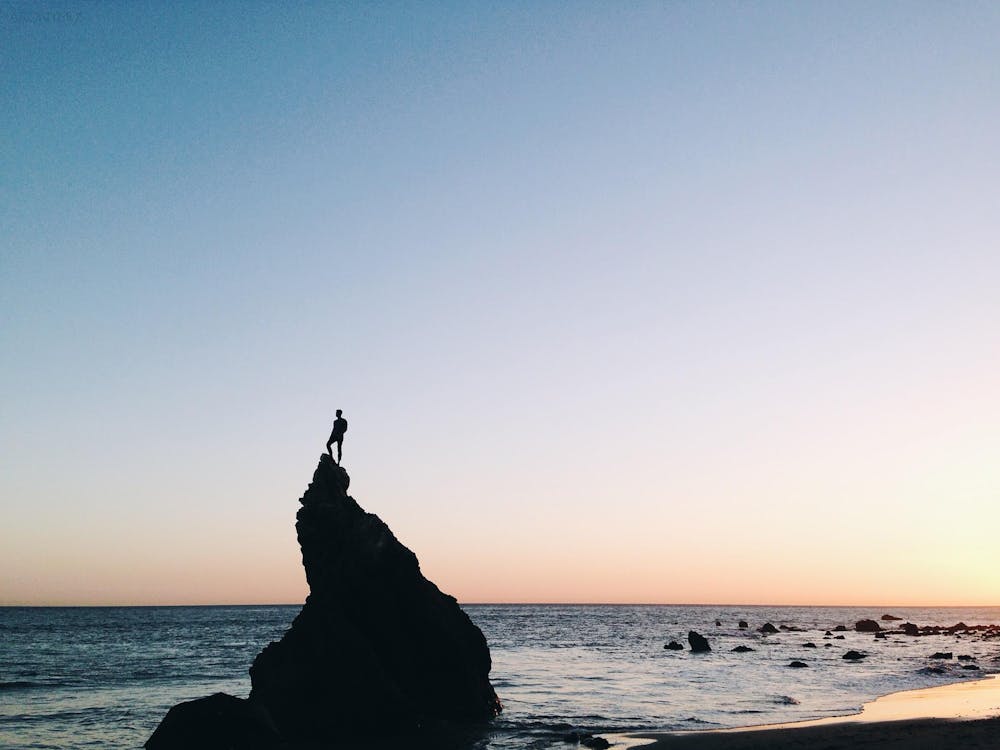 Silhouette of Person Standing on Rock on Beach Shore during Yellow Sunset