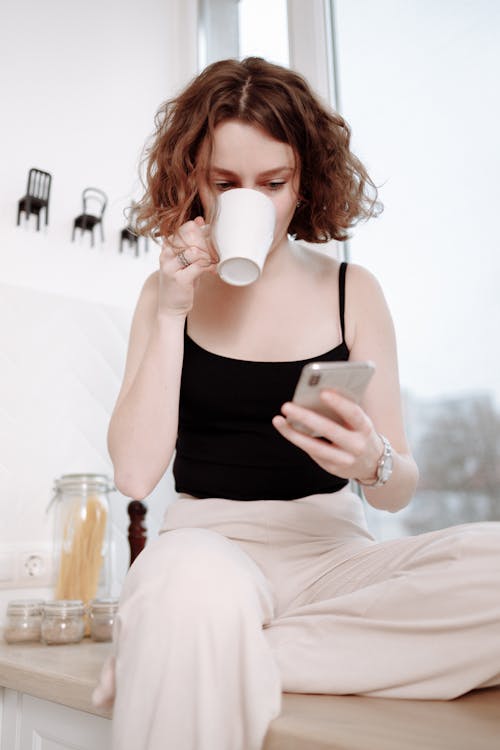 Woman Drinking from a Mug While Holding a Cellphone