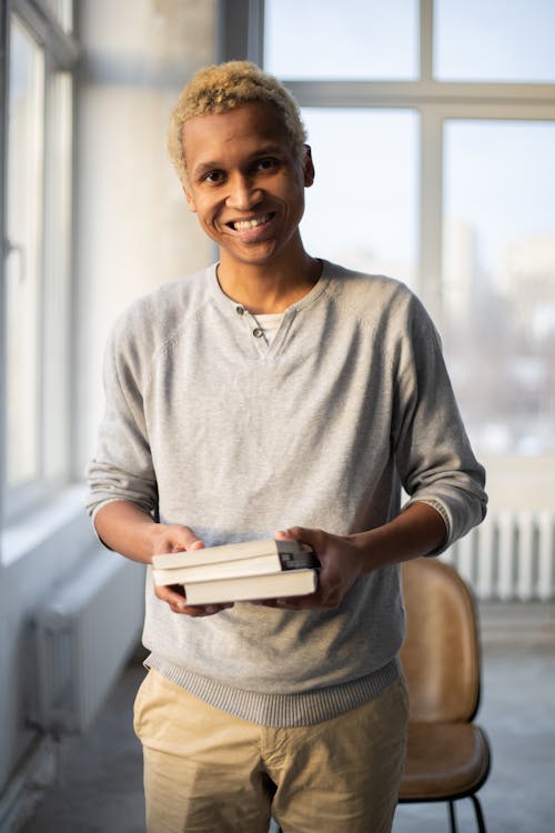 Cheerful African American male with dyed hair demonstrating books smiling and looking at camera near windows